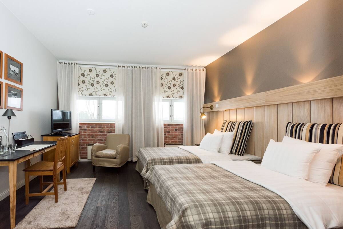 A hotel room with a desk, two beds, a TV, and a brick wall.