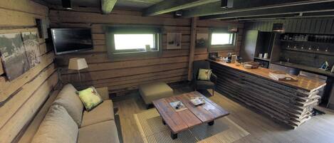 Una habitación acogedora de estilo cabaña, con detalles de madera, un sofá, una mesa de centro, un televisor montado en la pared y una zona de bar con mostrador y estanterías.