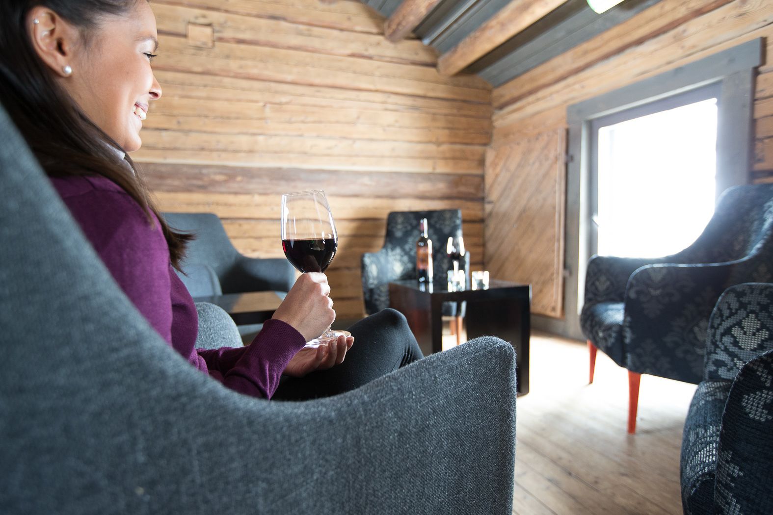 En person sidder på en grå sofa i et hyggeligt rum i træhusstil, med et glas rødvin, et lille bord med flasker og mønstrede lænestole.