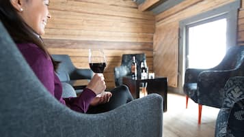 Una persona sentada en un sofá gris en una habitación estilo cabaña de madera, con una copa de vino tinto, una mesita baja con botellas y sillones estampados.