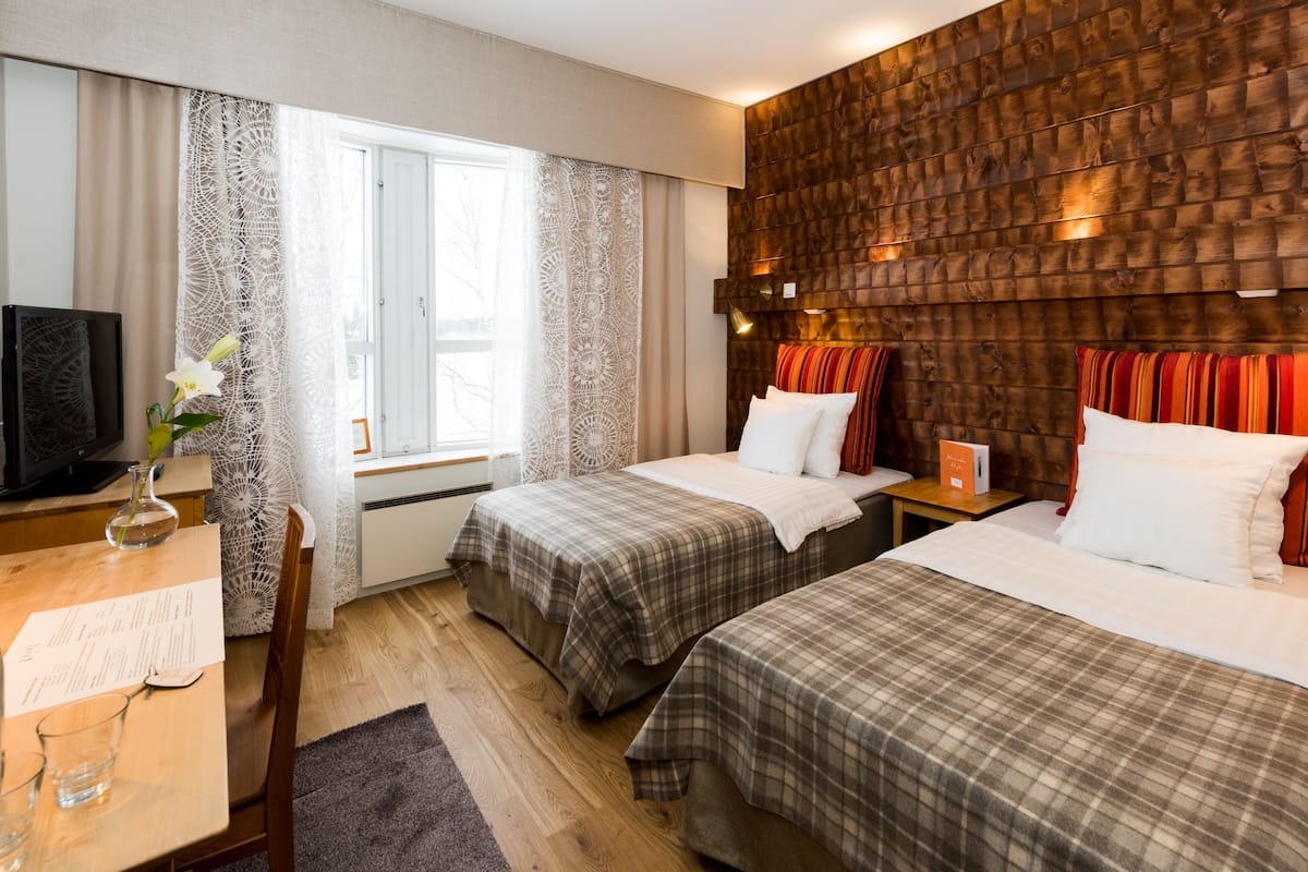 A hotel room with two beds, a wooden headboard, a desk with a chair, a TV, and a window with curtains.