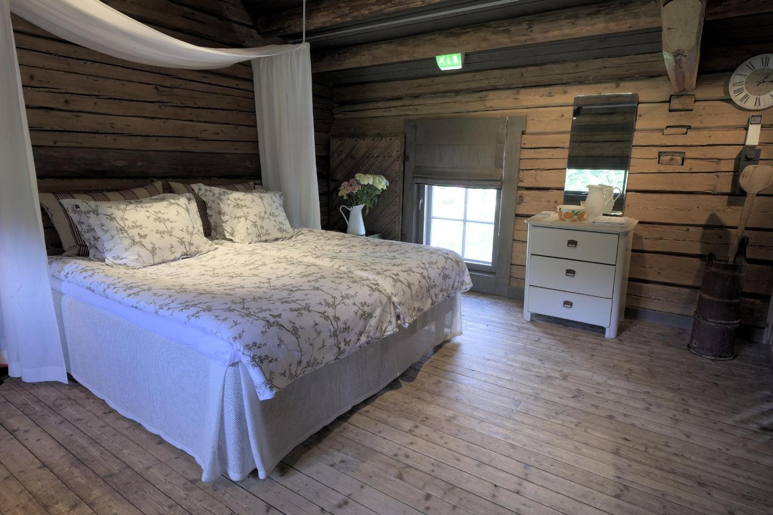 A wooden cabin bedroom with a bed, a nightstand, a window, and a clock on the wall.