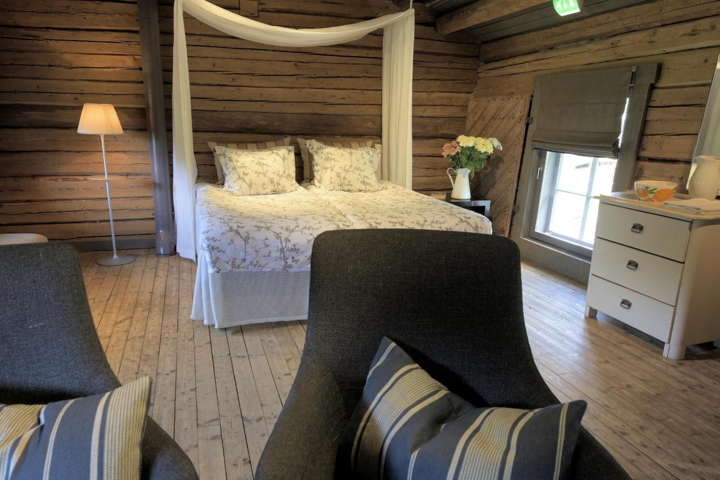 A wooden cabin bedroom with a canopy bed, a bedside table with a vase of flowers, and a dresser with a mirror.