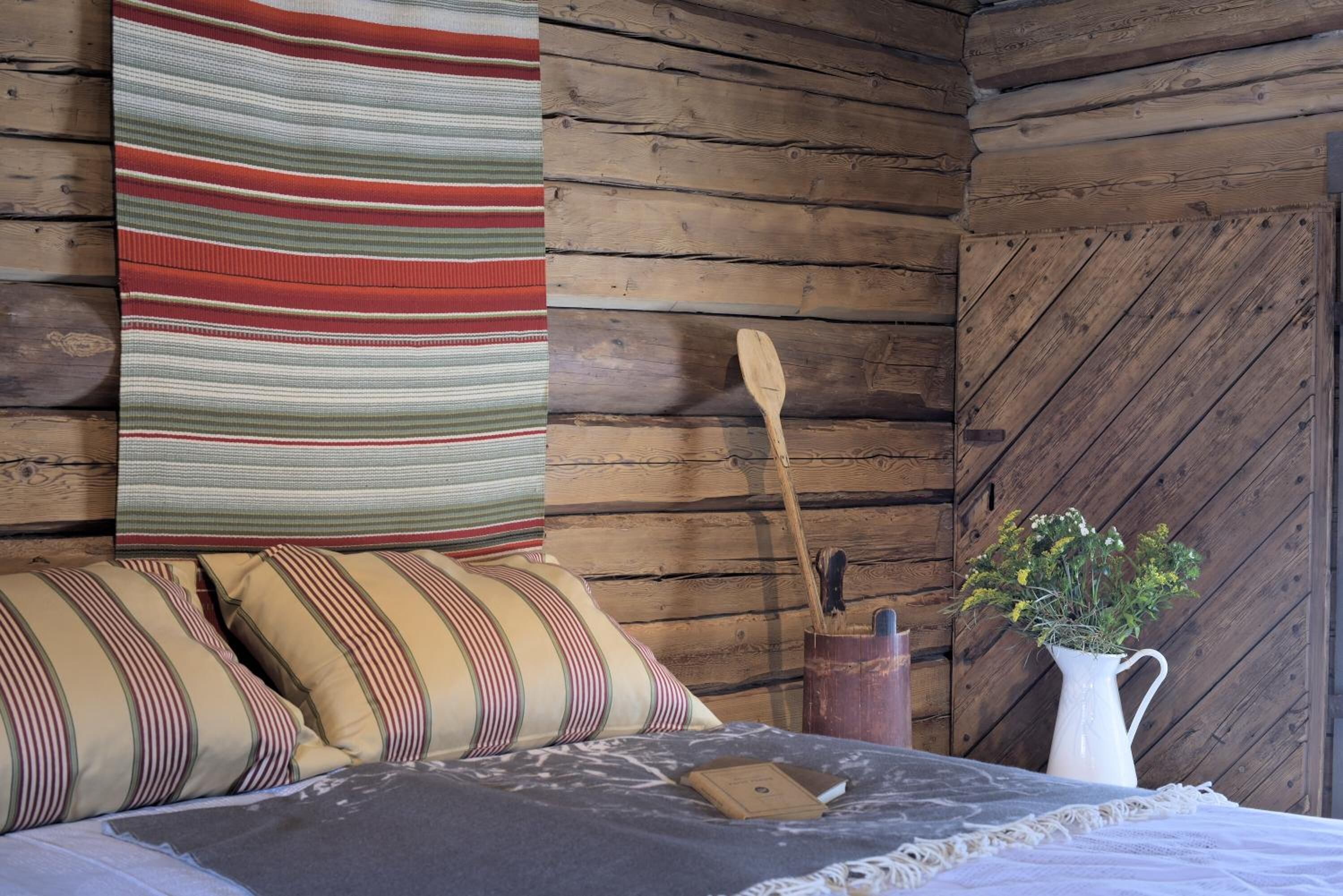 Une pièce chaleureuse avec un mur en bois brut, une couverture à rayures, des coussins à rayures, une cuillère en bois, un vase blanc rempli de fleurs et un livre posé sur le lit.