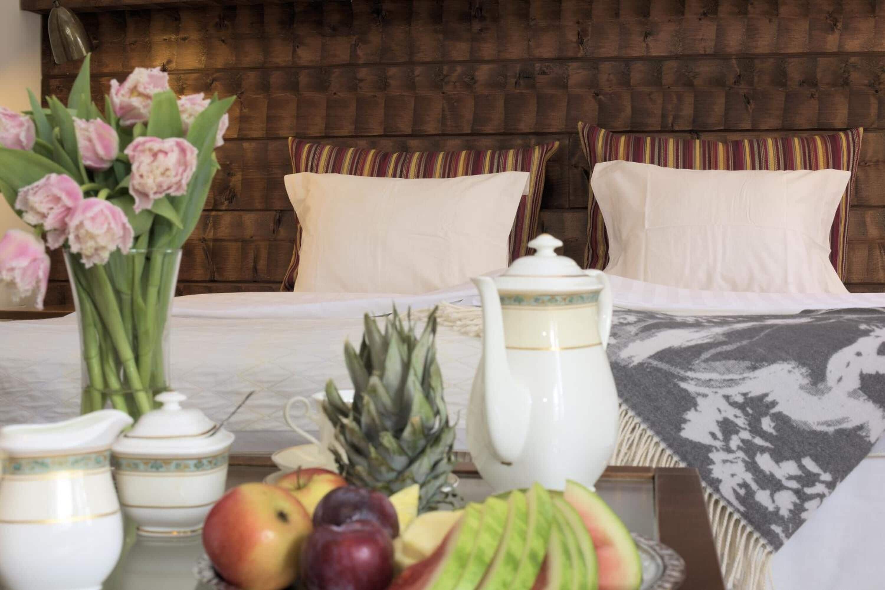 A neatly arranged hotel room with a bed, pillows, a vase of flowers, a fruit bowl, and a teapot.