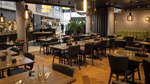 Un ristorante con tavoli e sedie in legno, lampade a sospensione e un'area salotto con cuscini verdi.