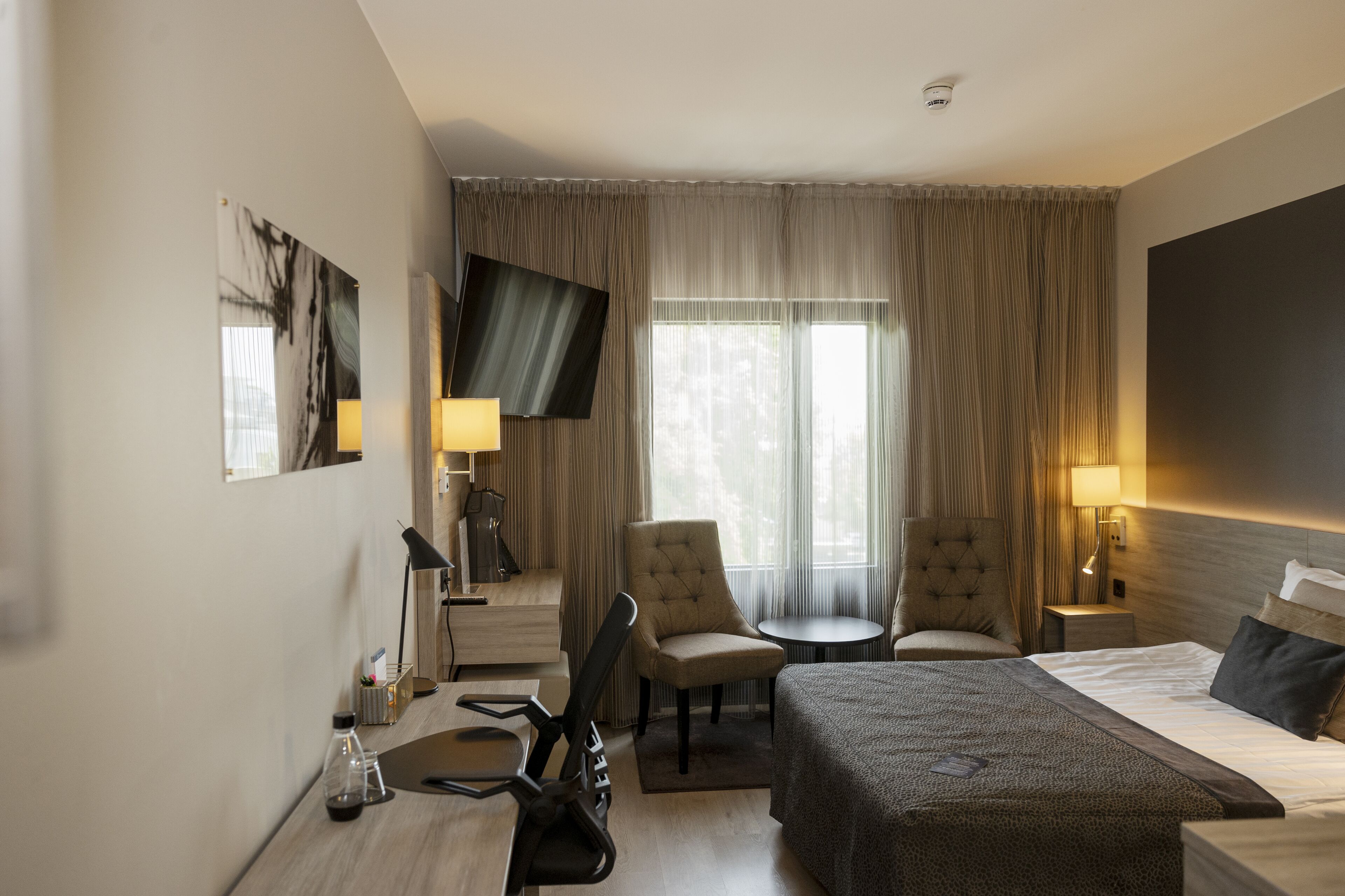 Een hotelkamer met een bed, twee fauteuils, een bureau met lamp, een televisie en een raam met gordijnen.