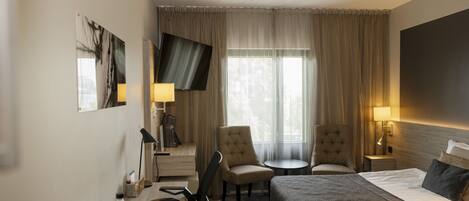 Habitación de hotel con una cama, dos sillones, un escritorio con lámpara, televisor y ventana con cortinas.