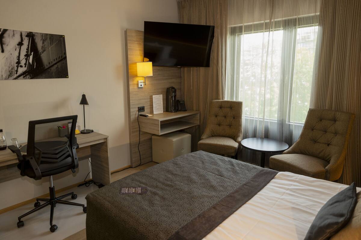 Une chambre d’hôtel comprenant un lit, deux fauteuils, un bureau avec un ordinateur, une télévision et une fenêtre avec des rideaux.