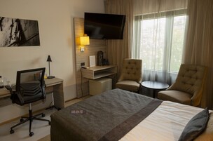 A hotel room with a bed, two armchairs, a desk with a computer, a television, and a window with curtains.