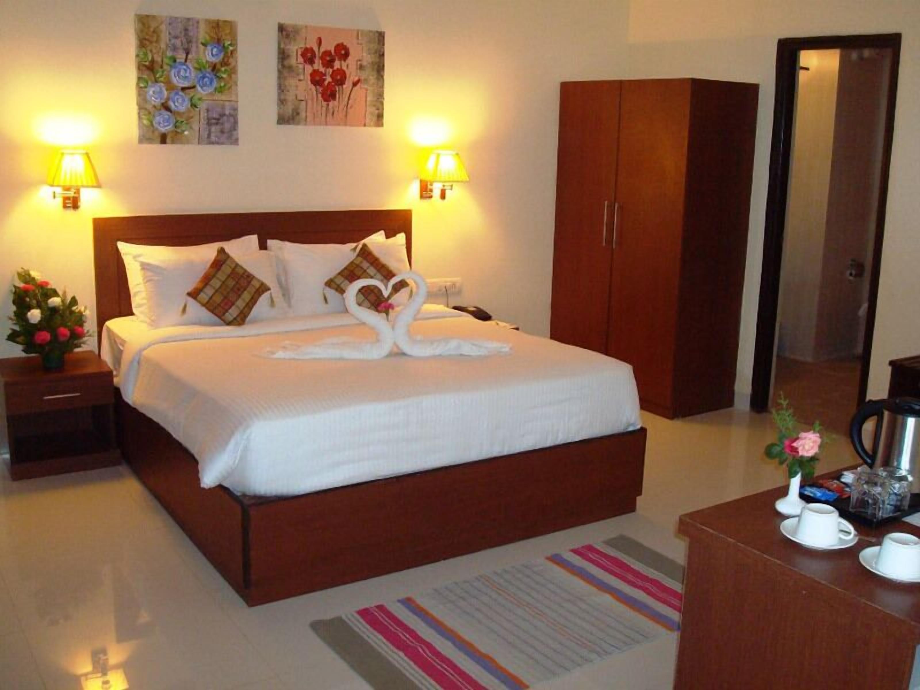 A neatly made bed with white linens and decorative pillows, a wooden wardrobe, a bedside table with a lamp, and a coffee station with cups and a kettle.