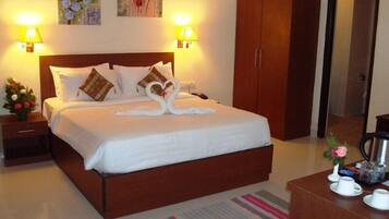 A neatly made bed with white linens and decorative pillows, a wooden wardrobe, a bedside table with a lamp, and a coffee station with cups and a kettle.