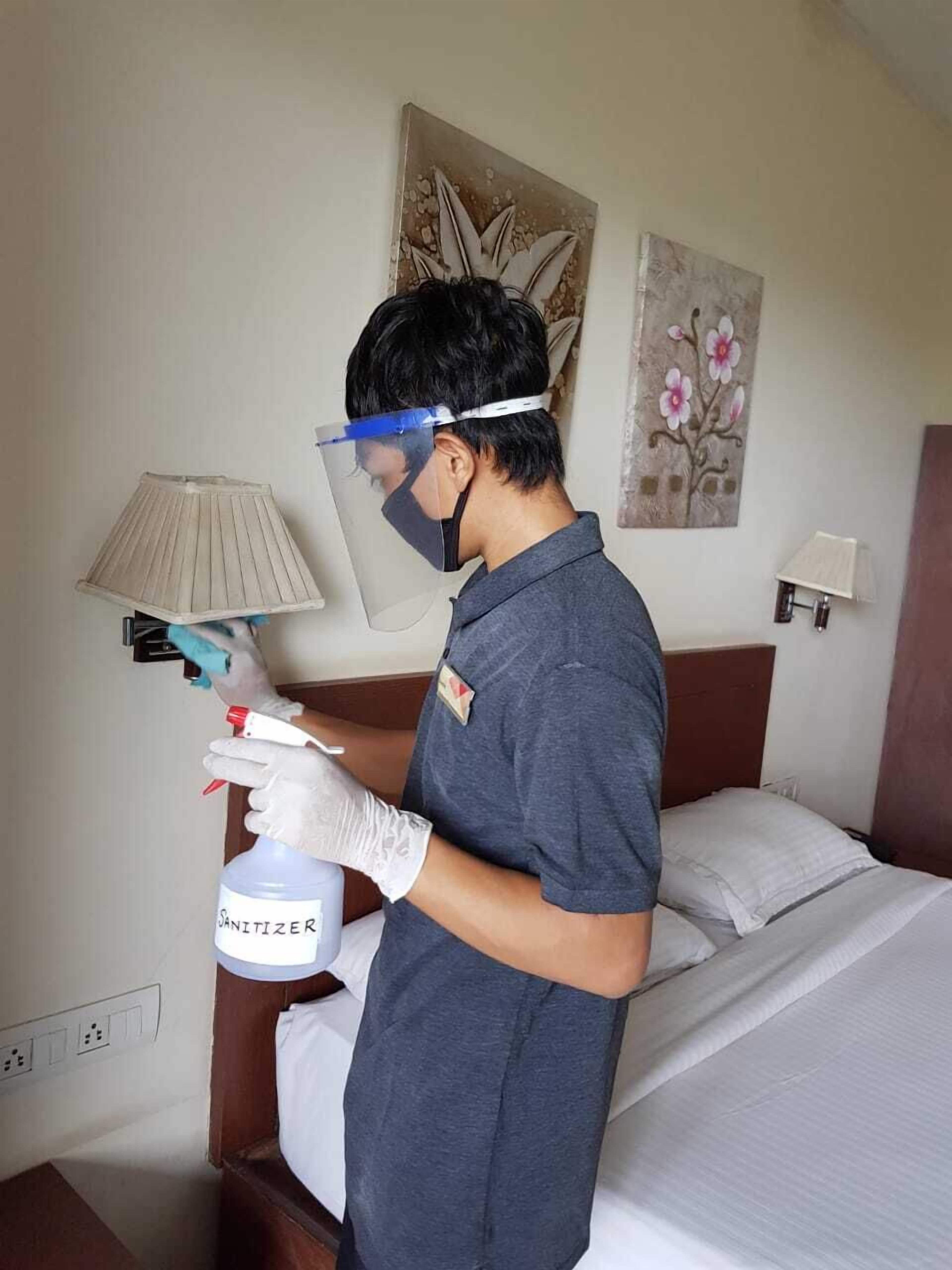 A person in a grey uniform spraying sanitizer in a hotel room with a bed, nightstand, lamp, and floral artwork on the wall.