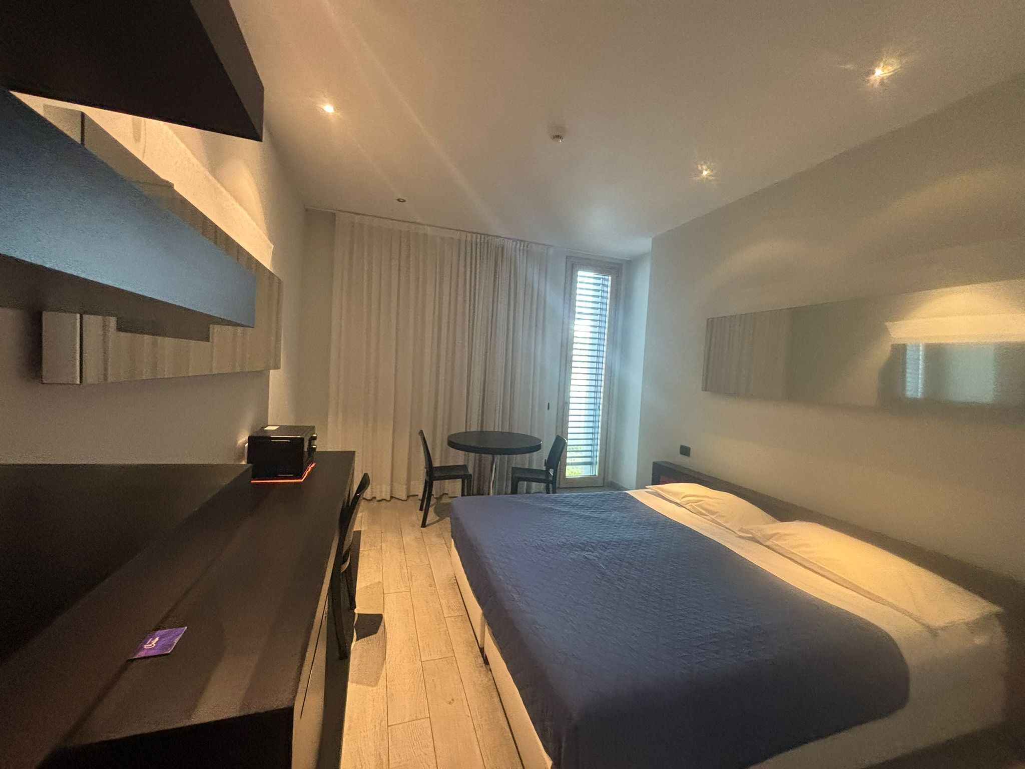 A modern hotel room with a bed, a desk with a computer, a chair, and a window with blinds.
