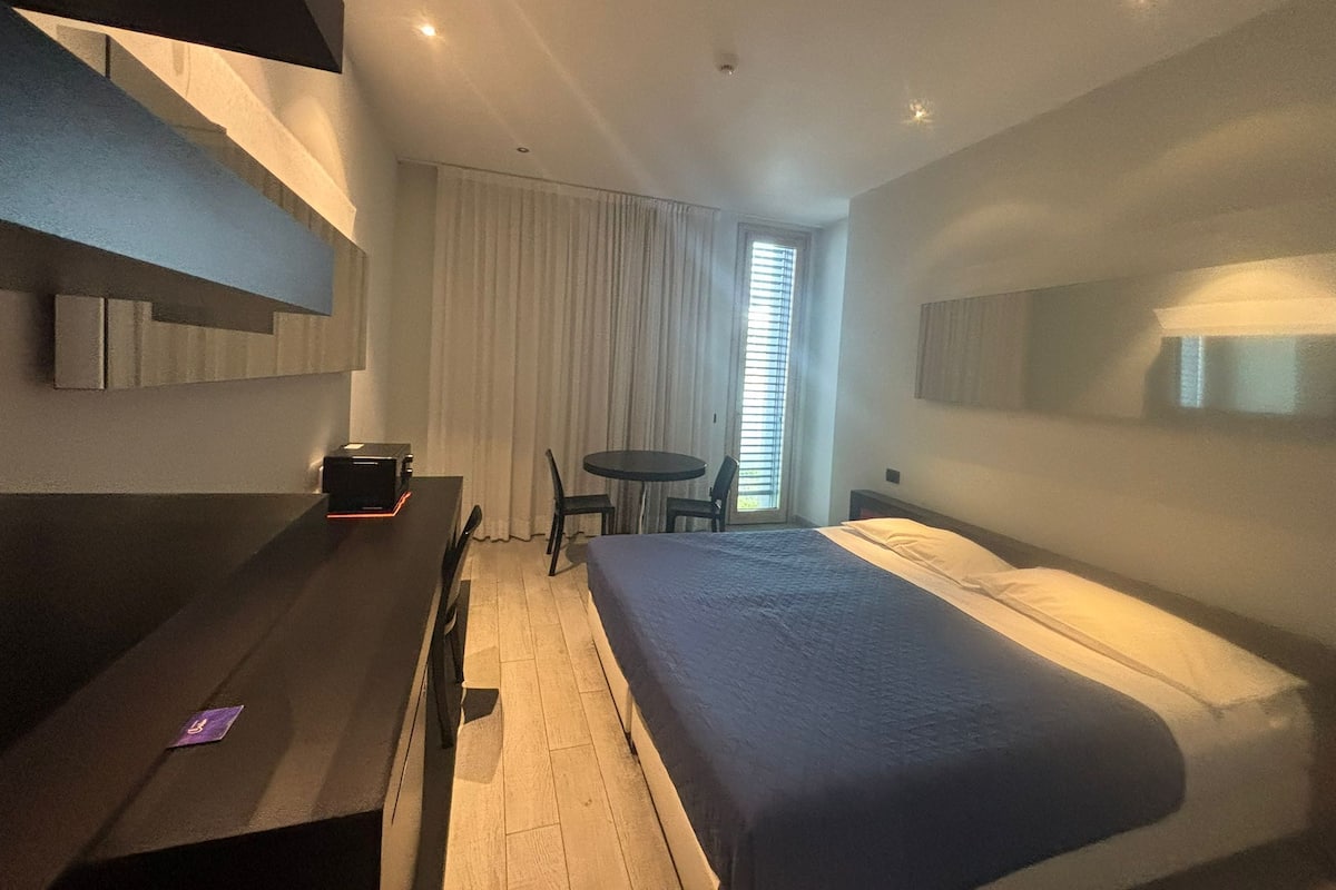 A modern hotel room with a bed, a desk with a computer, a chair, and a window with blinds.
