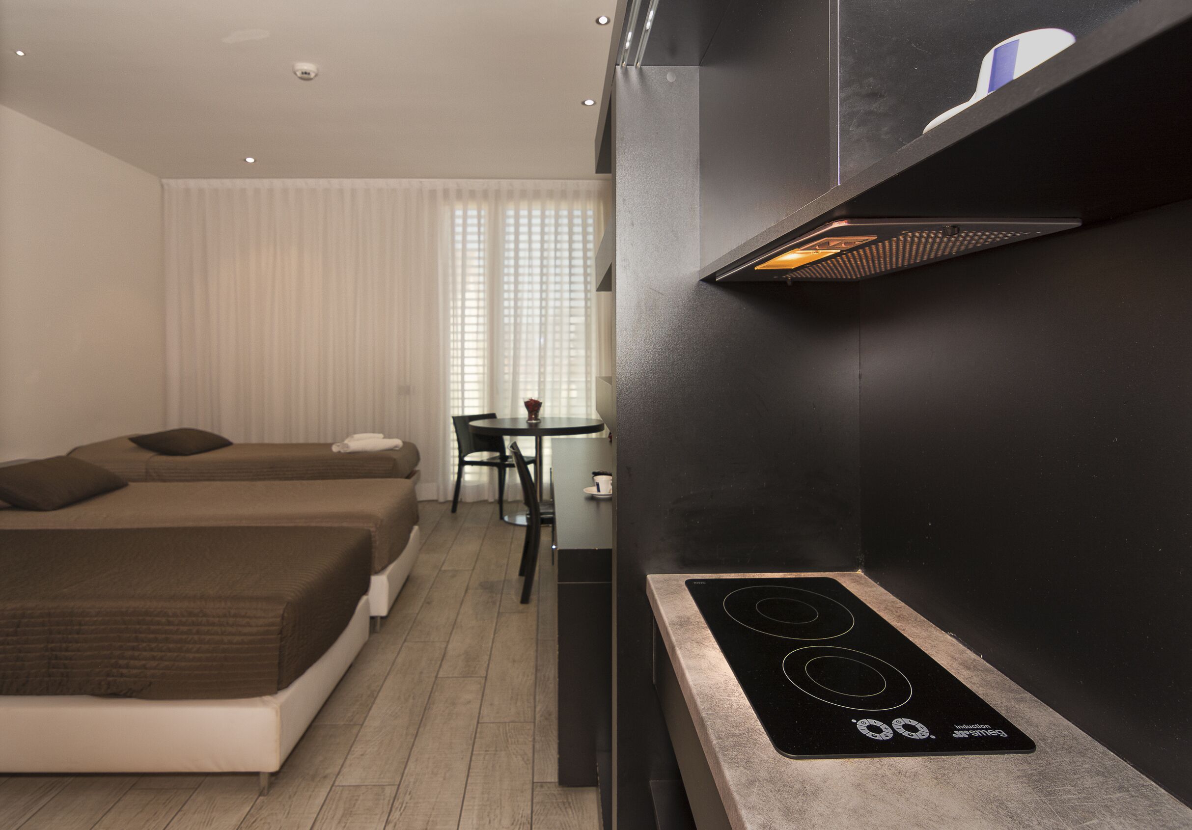 Ein modernes Hotelzimmer mit Küchenzeile, zwei Betten, einem Esstisch und einem Fenster mit Jalousien.