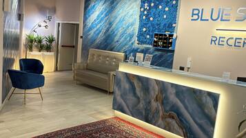 Una zona de recepción con pared de mármol, un sofá, una silla azul y una alfombra roja estampada.