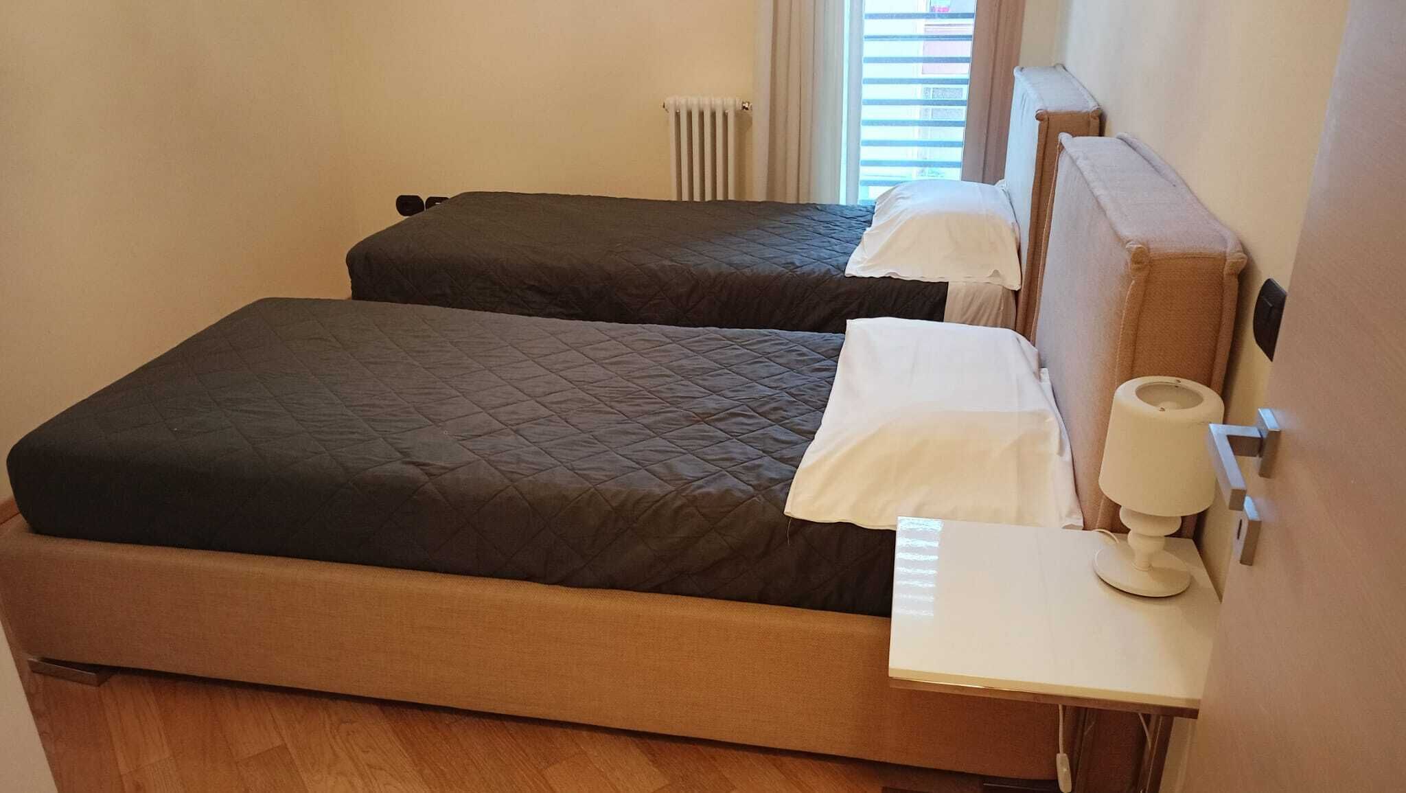 A hotel room with two beds, a bedside table with a lamp, and a radiator.