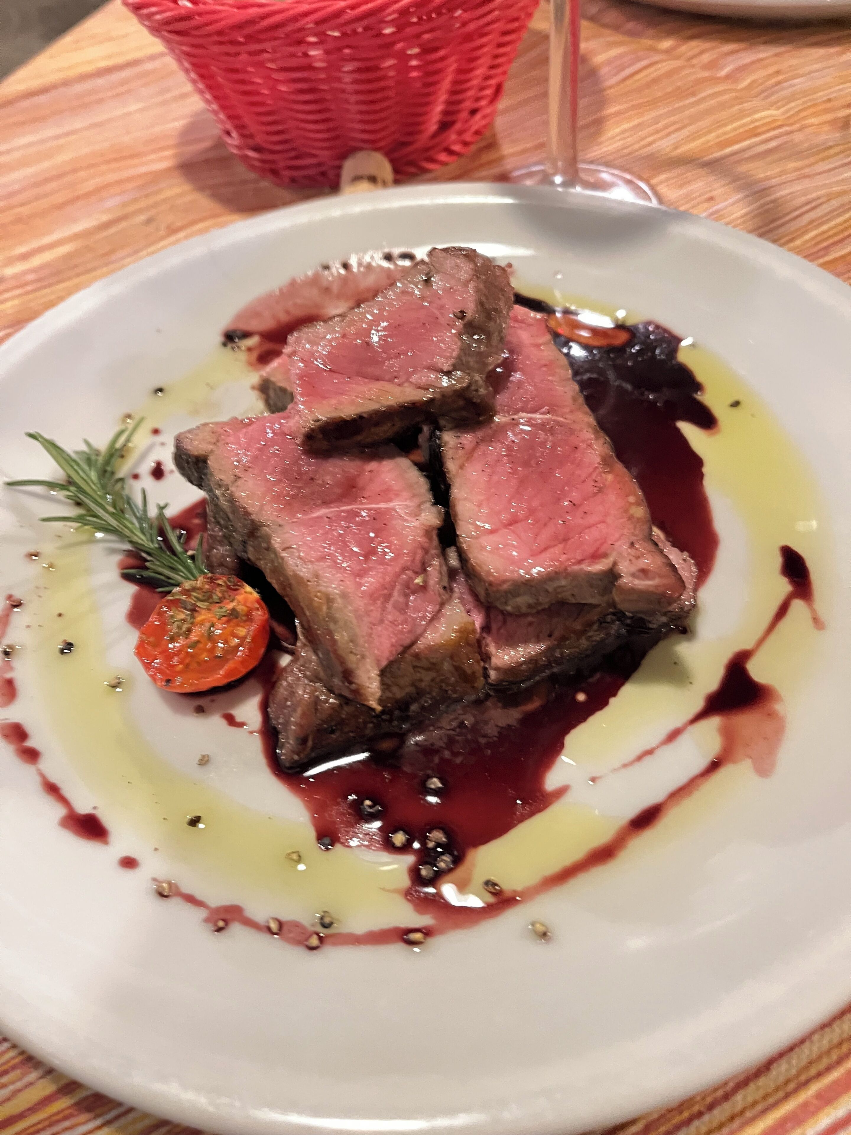 Un piatto di bistecca guarnito con rosmarino e un pomodorino ciliegino, servito su un piatto bianco con salsa rossa e un filo d'olio.