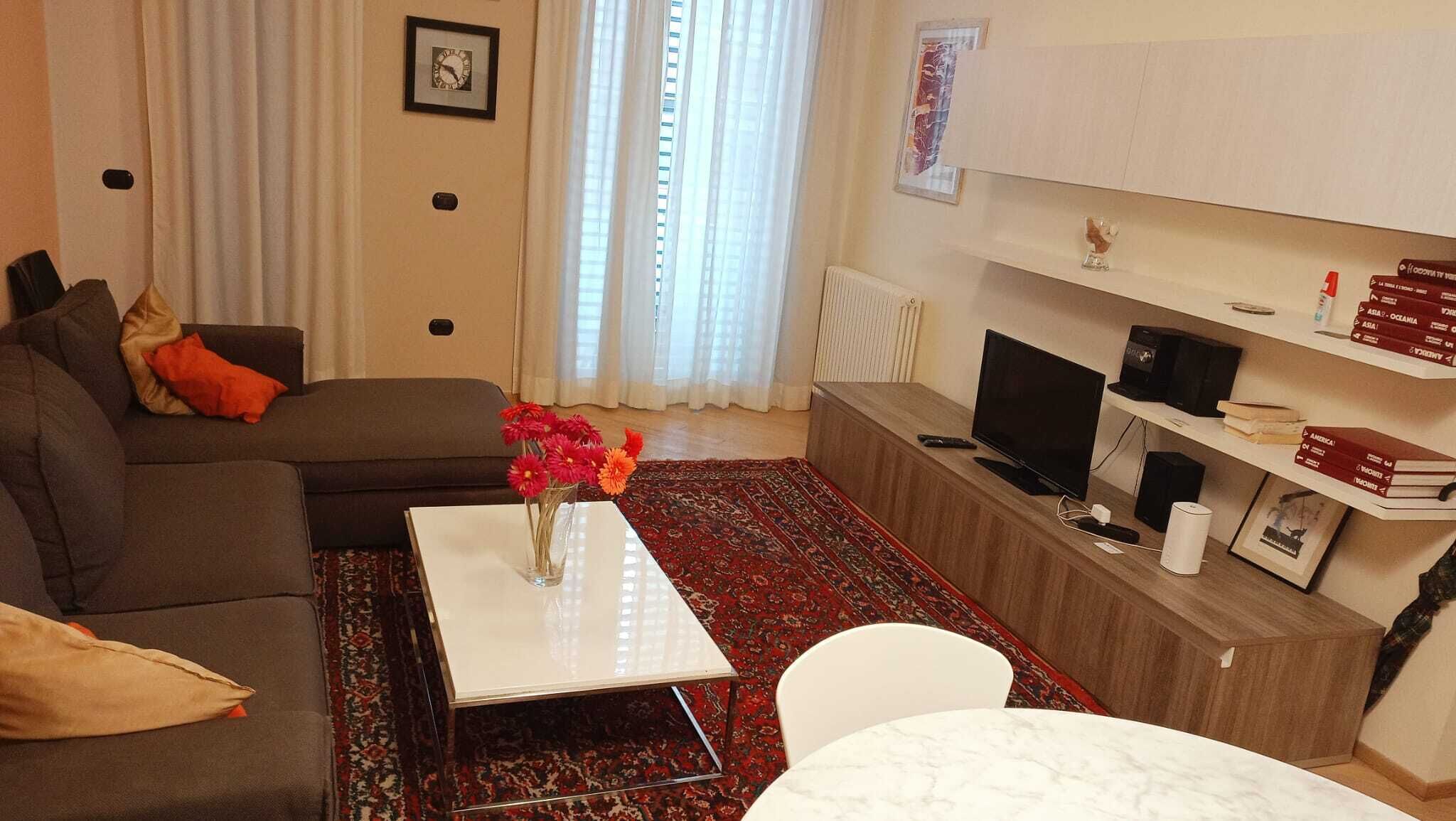 Una sala de estar con un sofá, una mesa de centro con flores, un televisor y estanterías.