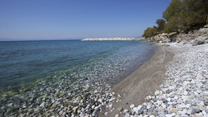 A pebbled beach with clear water, a rocky outcrop, and trees in the background. - Apollon Apartments (West Mani)