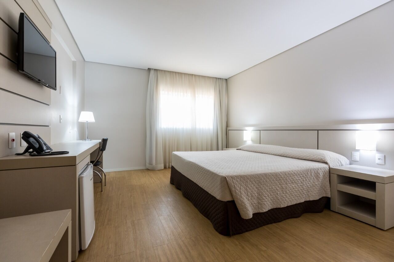A hotel room with a bed, bedside tables, a desk with a phone, a TV mounted on the wall, and a window with curtains.