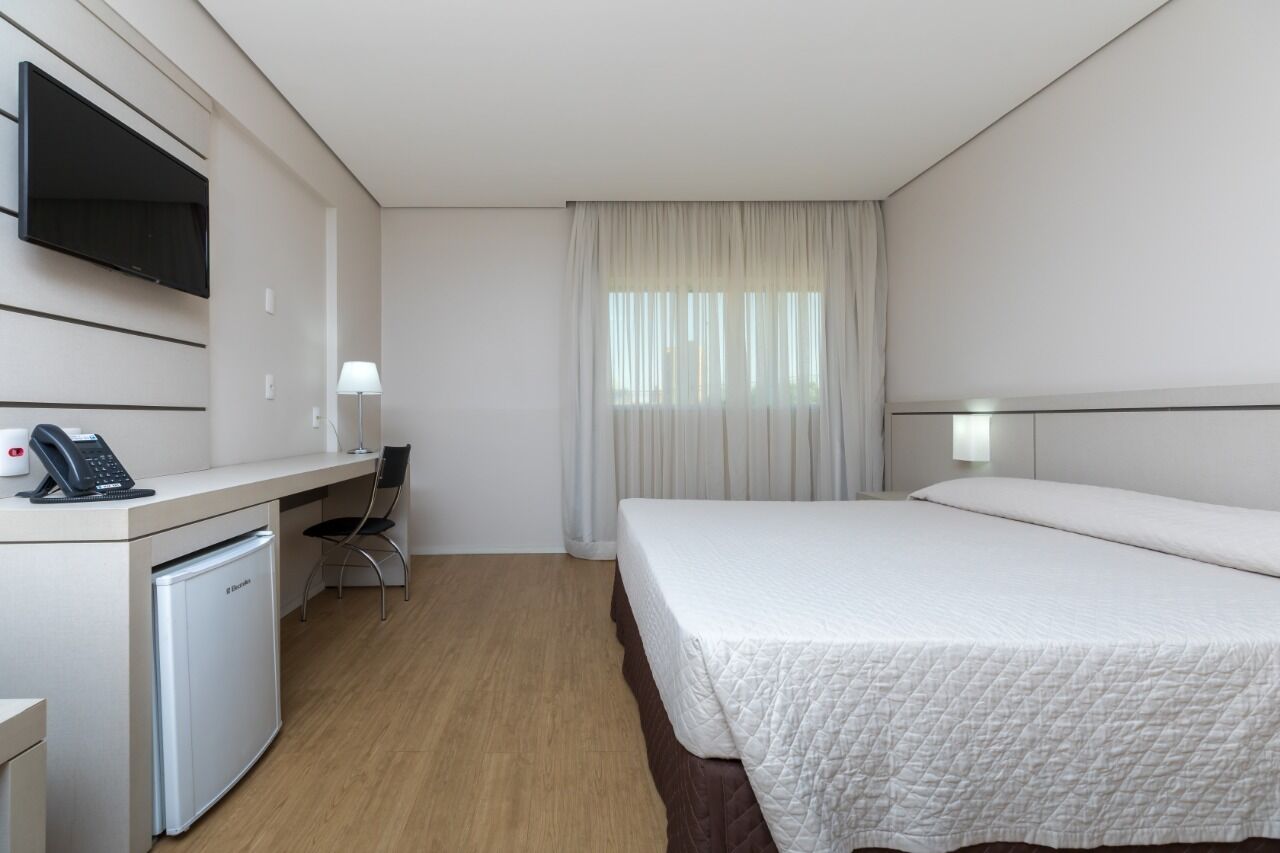 A hotel room with a bed, a desk with a phone, a small refrigerator, a chair, and a TV mounted on the wall.