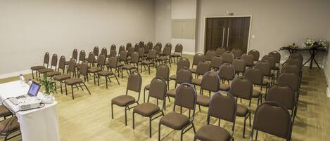 A conference room with rows of brown chairs, a white table with a laptop and water bottles, and a projector on the ceiling.