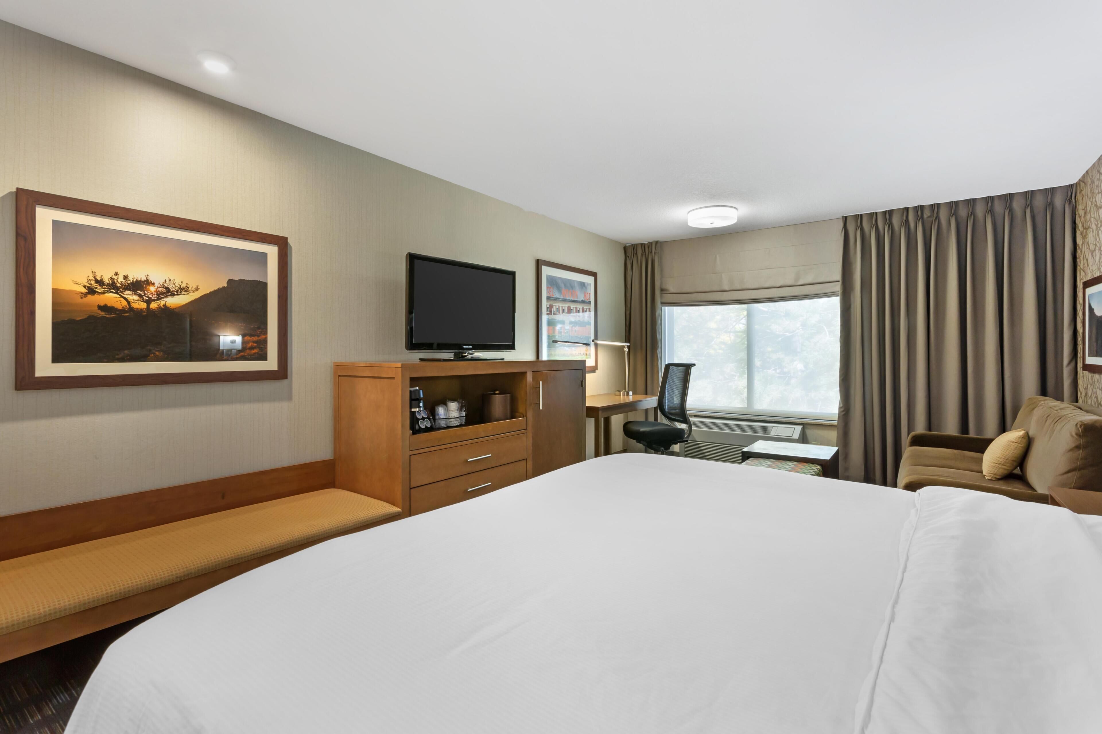 A hotel room with a large bed, a desk with a chair, a flat-screen TV, a framed picture, and a window with curtains.