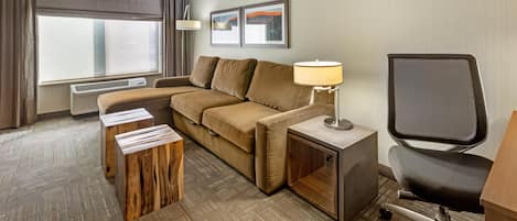 Habitación de hotel con sofá, mesita de madera, lámpara, silla y un cuadro en la pared.