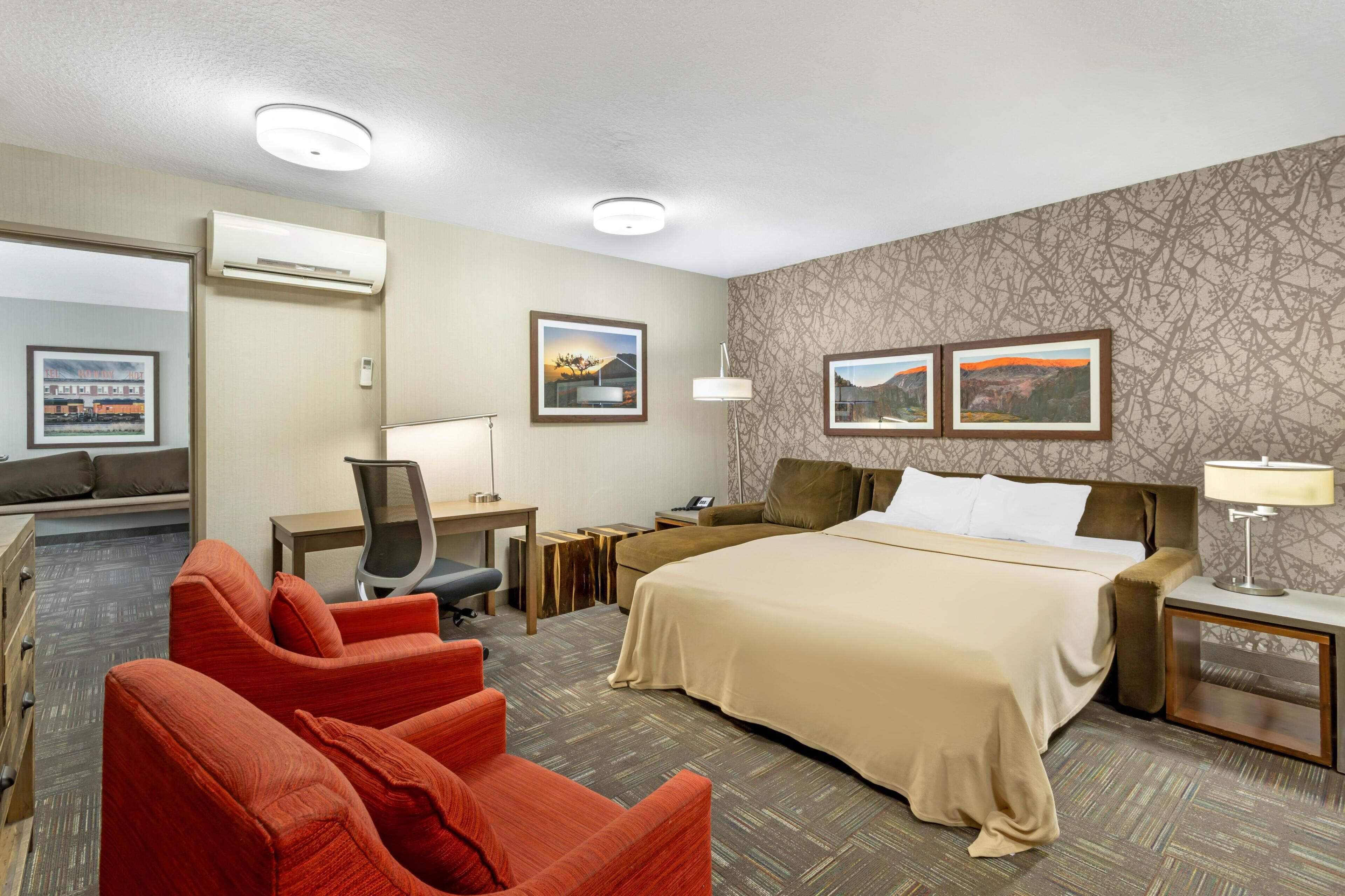 Quarto de hotel com uma cama, duas poltronas vermelhas, uma escrivaninha com cadeira, uma mesa de cabeceira, um abajur e duas imagens emolduradas na parede.
