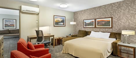 Ein Hotelzimmer mit einem Bett, zwei roten Sesseln, einem Schreibtisch mit Stuhl, einem Nachttisch, einer Lampe und zwei gerahmten Bildern an der Wand.