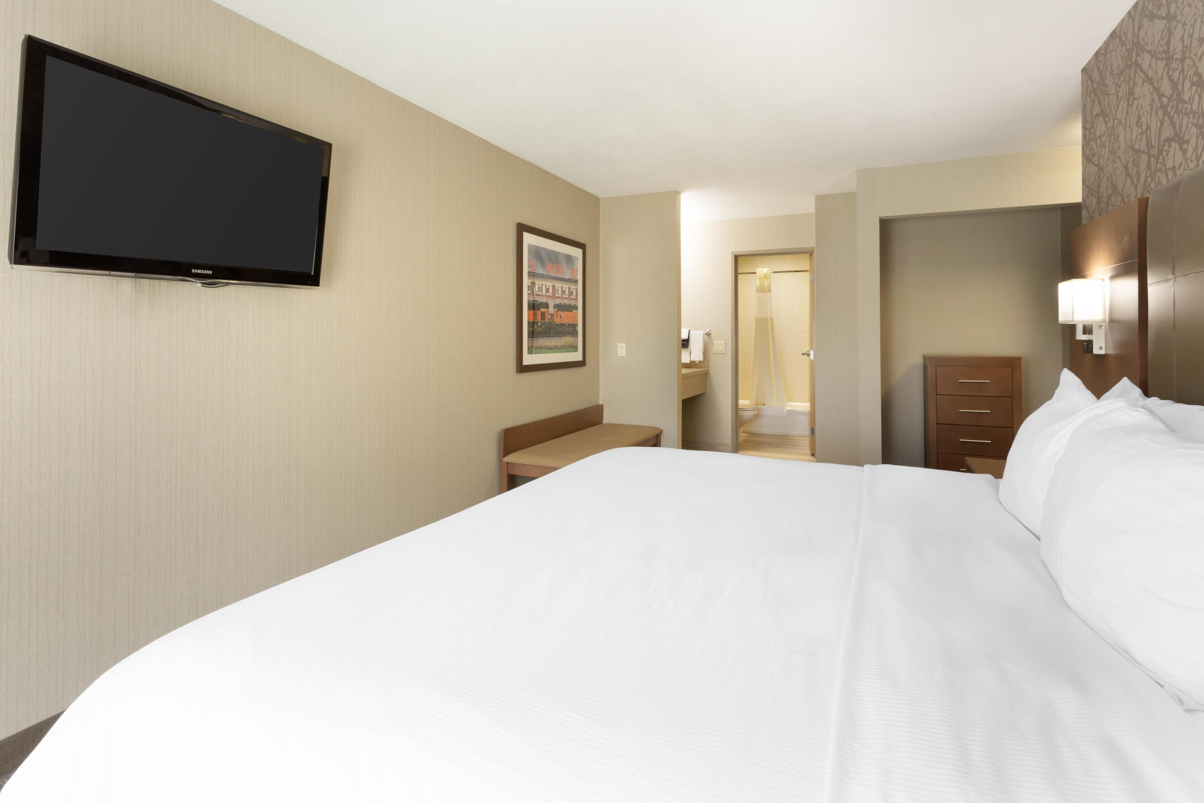 Une chambre d’hôtel avec un grand lit, un téléviseur à écran plat fixé au mur, un tableau encadré, un banc, une commode et une porte donnant sur une salle de bain.