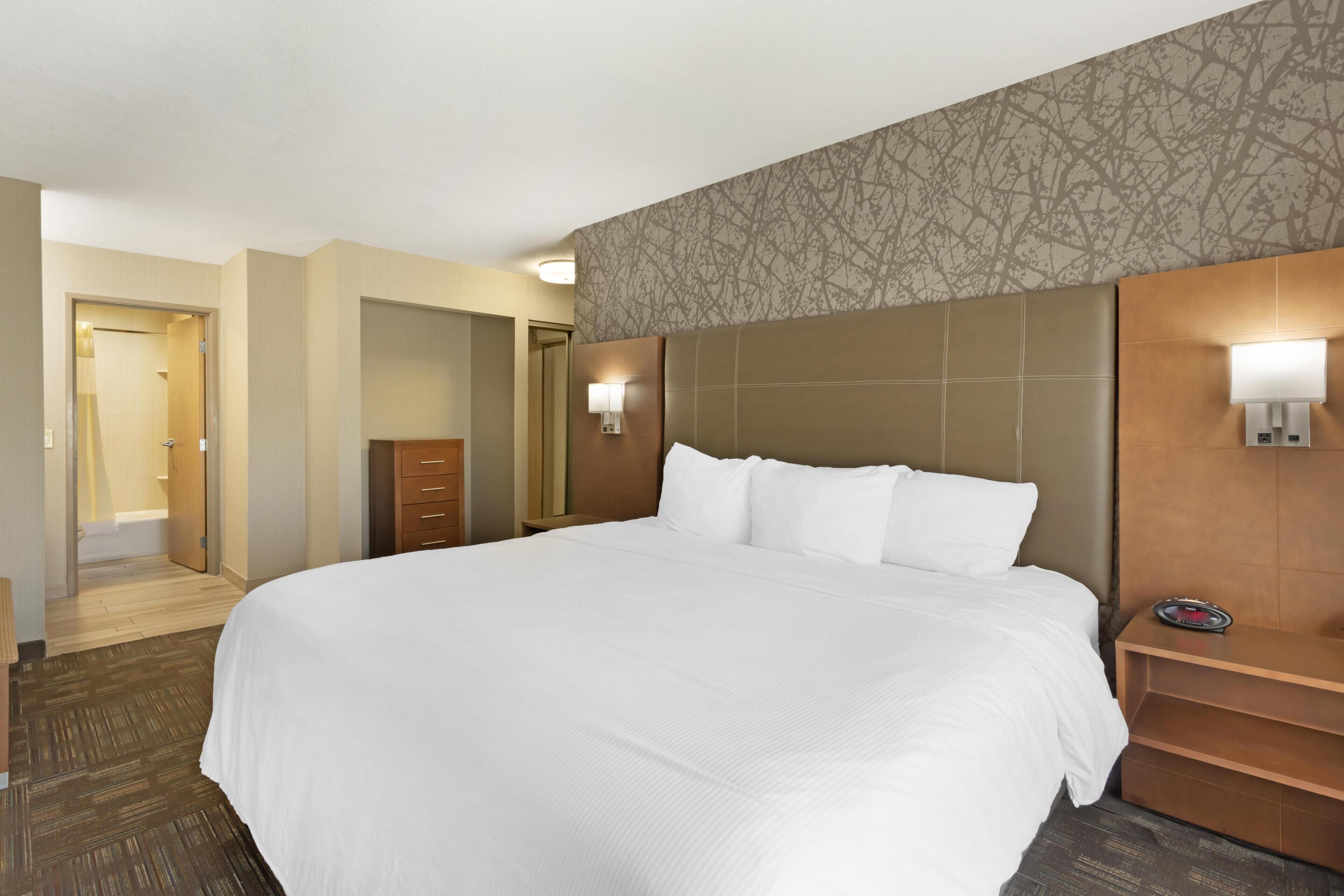 Une chambre d’hôtel avec un grand lit, une commode en bois, une table de chevet avec une lampe et une salle de bain visible à travers une porte ouverte.
