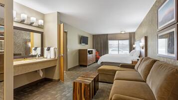 Ein Hotelzimmer mit einem Bett, einer Couch, einem Holztisch, einem Fernseher und einem Badezimmer mit Spiegel und Handtuchhalter.