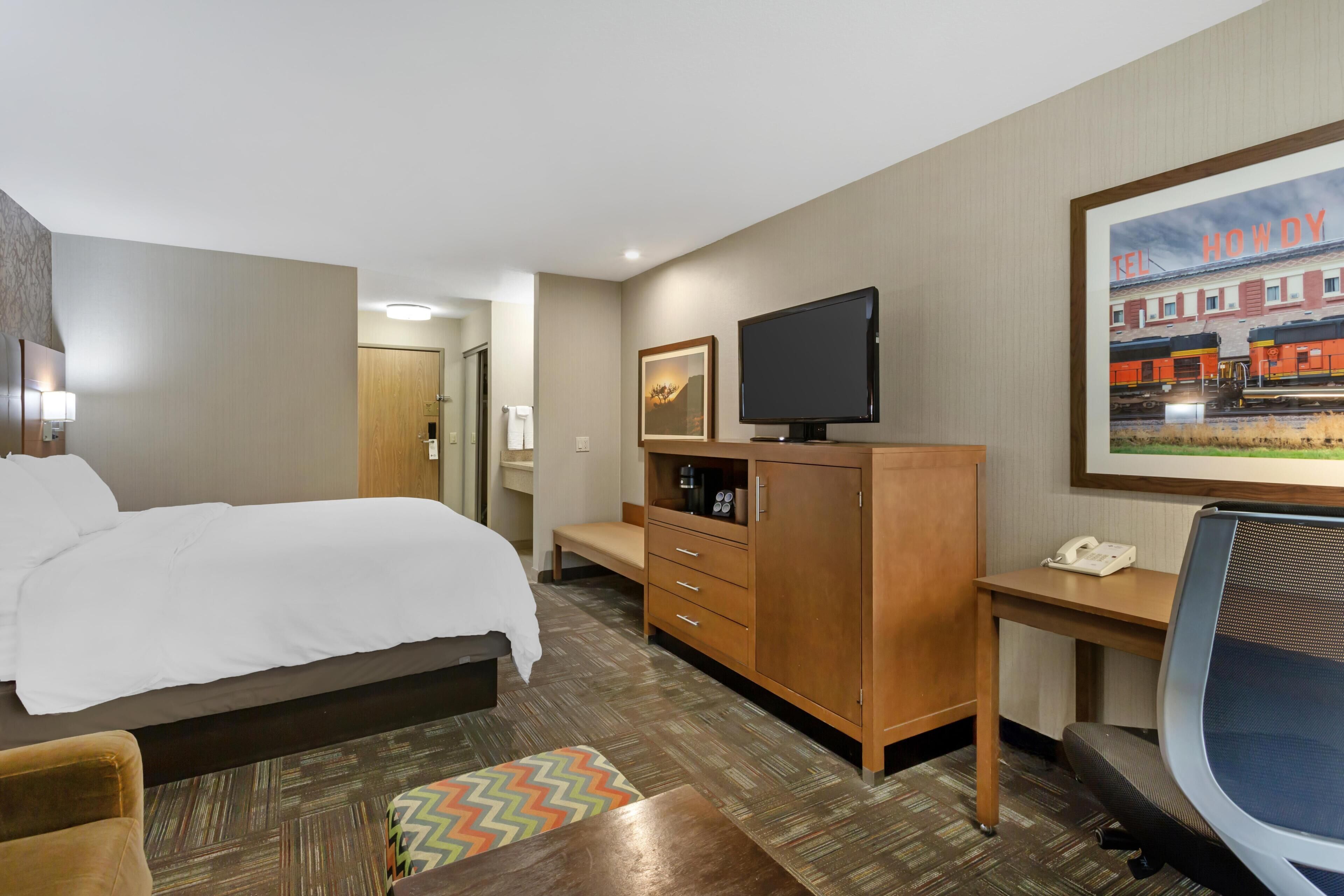 A hotel room with a bed, a desk, a television, a telephone, and a painting of a train on the wall.