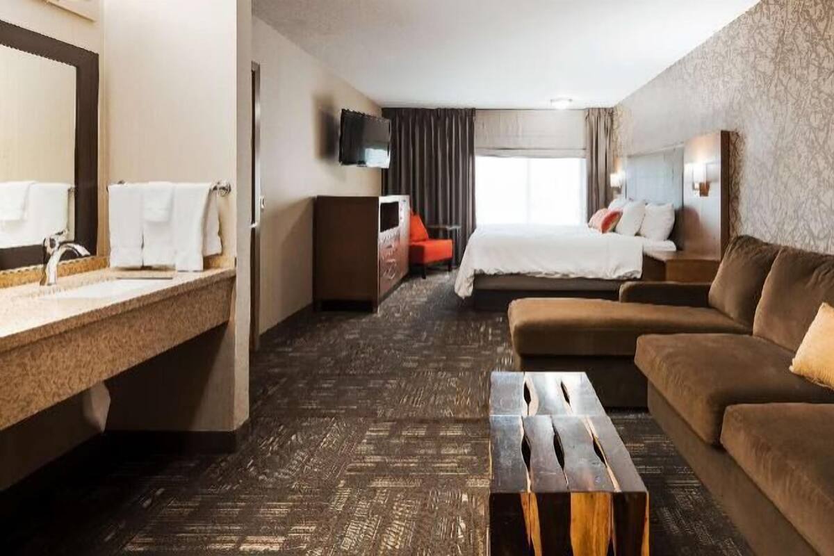 Habitación de hotel con cama, sofá, mesa de centro, televisor y baño con lavamanos y espejo.