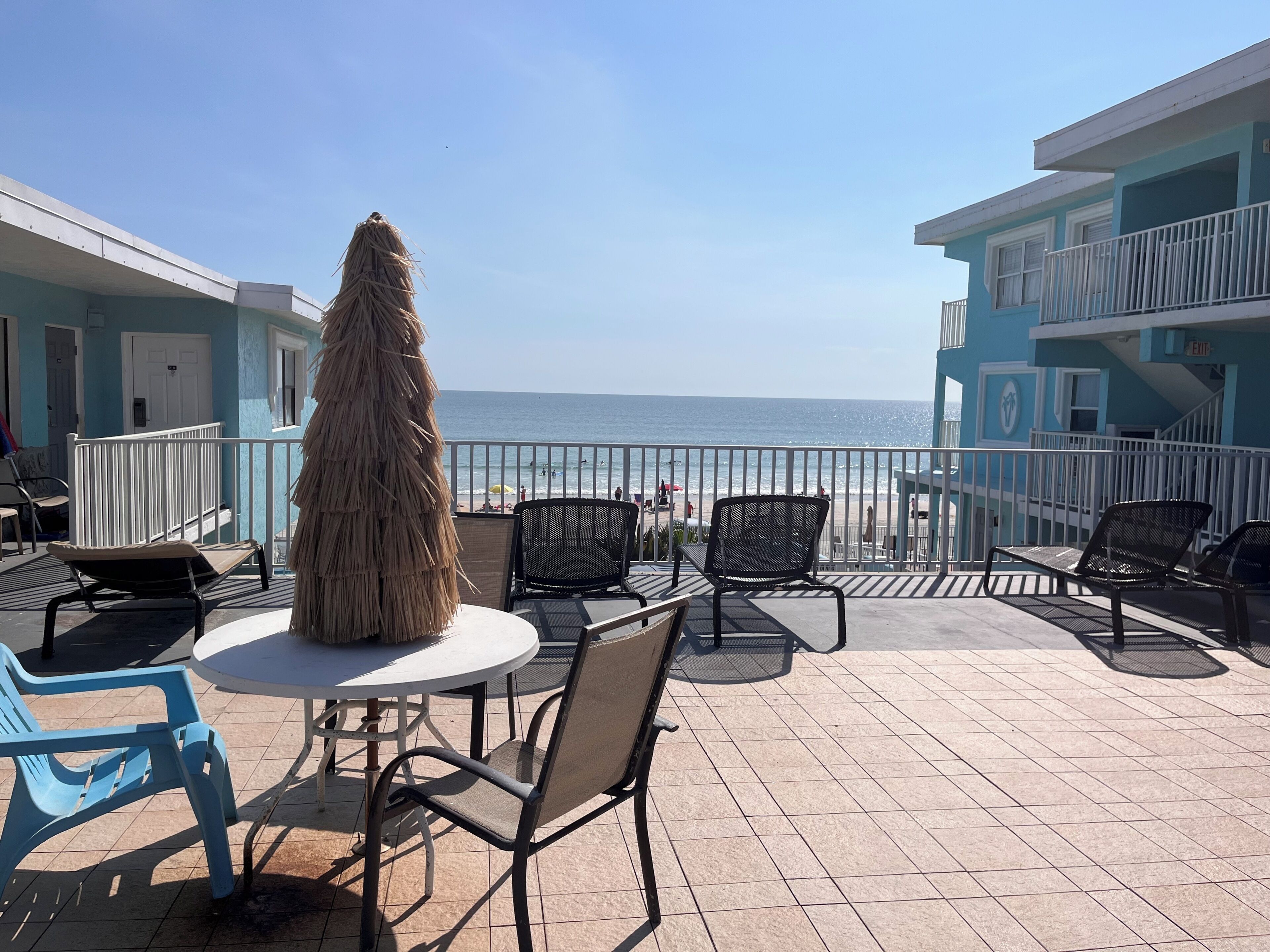 An outdoor seating area with a view of the ocean, featuring a table and chairs, a thatched umbrella, and a building with a balcony.