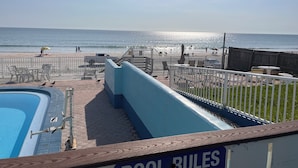 Area piscina con cartello che indica le regole, terrazza recintata con sedie e tavoli, e vista sulla spiaggia e sull'oceano.