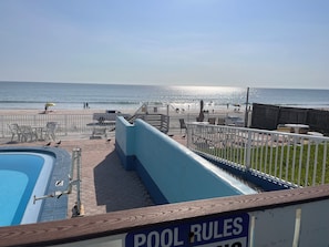A pool area with a sign indicating pool rules, a fenced deck with chairs and tables, and a view of the beach and ocean. - Sand Castle Motel (Daytona Beach Shores)