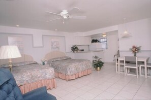 A bedroom with two beds, a ceiling fan, a desk with chairs, and a small kitchen area.