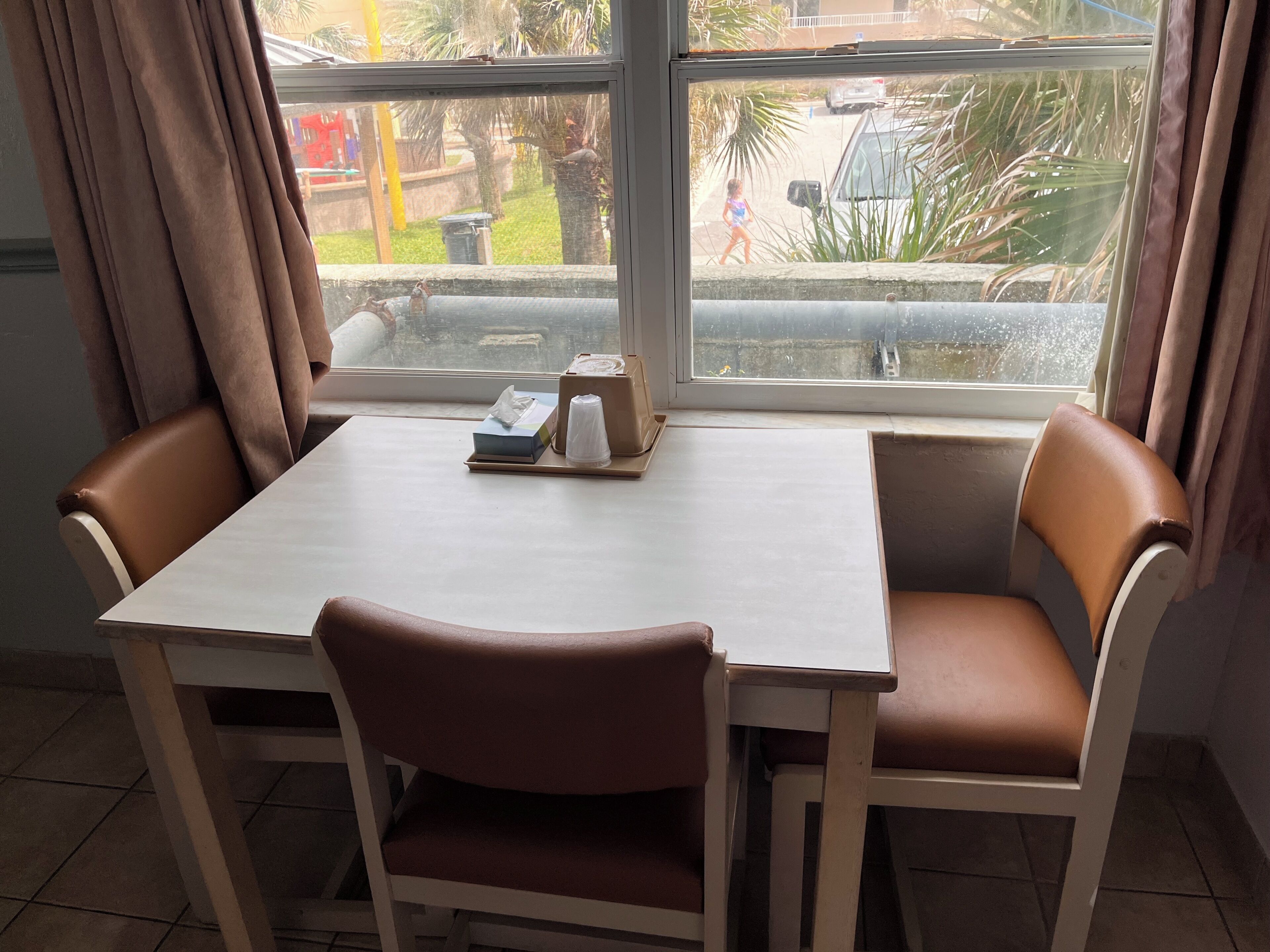 Uma sala de jantar com uma mesa e cadeiras, uma janela com vista para o exterior, e uma pequena mesa com um estojo de lenços de papel e uma chávena.