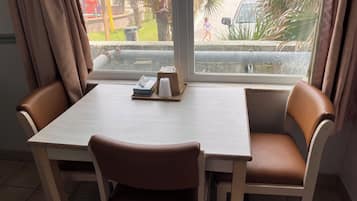 Uma sala de jantar com uma mesa e cadeiras, uma janela com vista para o exterior, e uma pequena mesa com um estojo de lenços de papel e uma chávena.