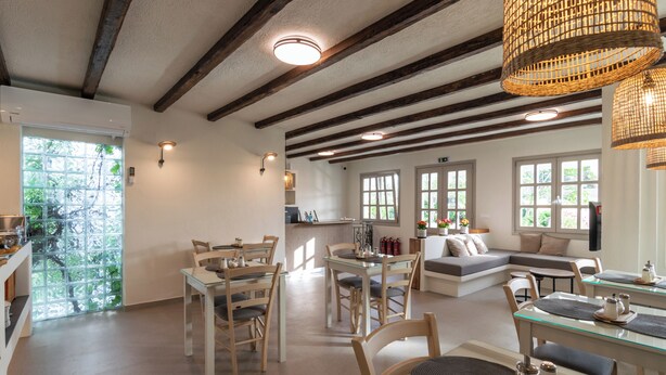 A spacious living area with a dining table, chairs, and a sofa. There is a kitchen area in the background with a window. - Hotel Rigas (Milos)