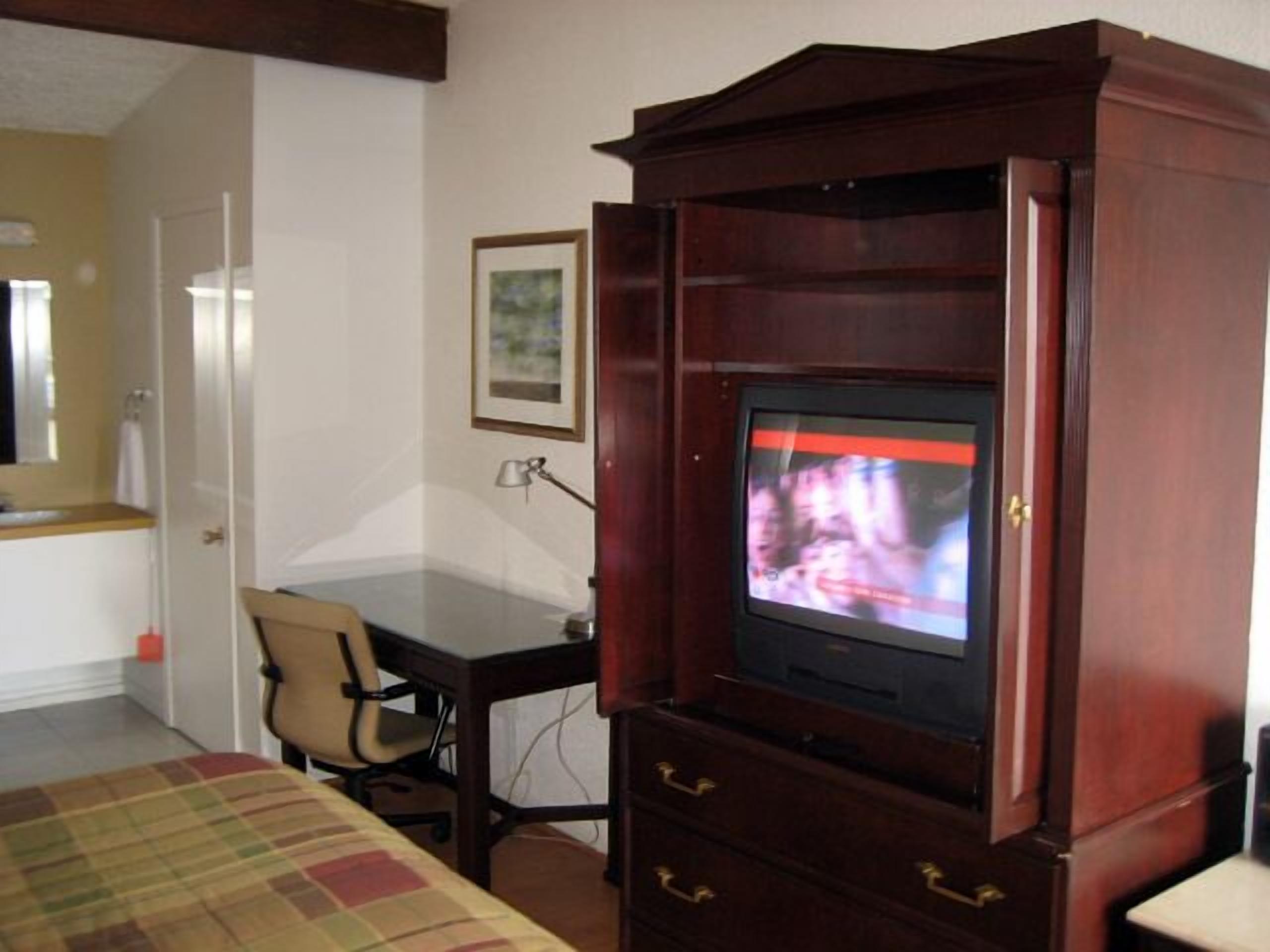 Une chambre d’hôtel avec un meuble de rangement en bois servant de centre de divertissement, un lit recouvert d’une couverture à carreaux, un bureau avec une chaise et un tableau encadré accroché au mur.