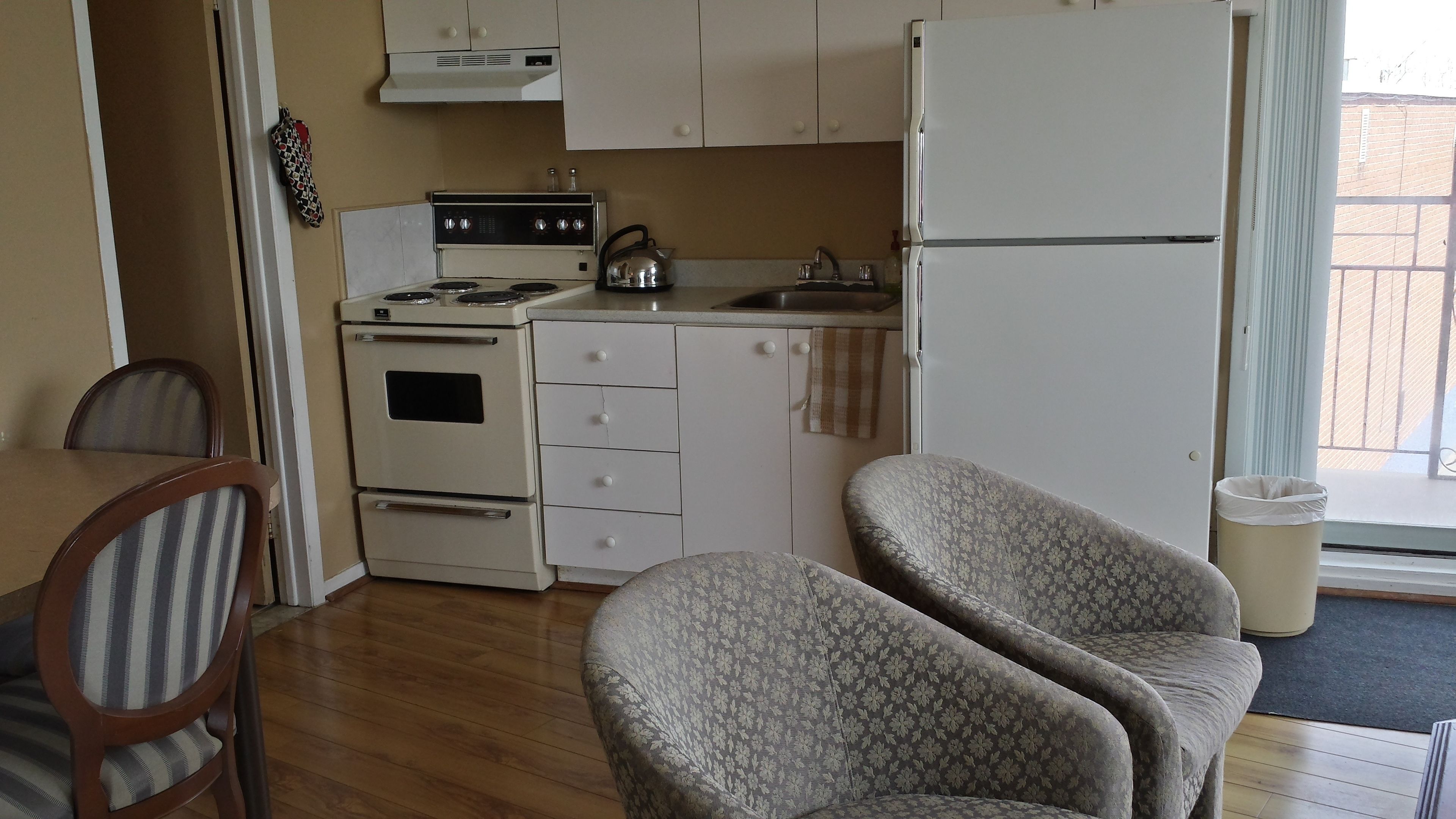 Una cocina compacta con estufa, pileta y refrigerador, sillas de madera y un bote de basura.