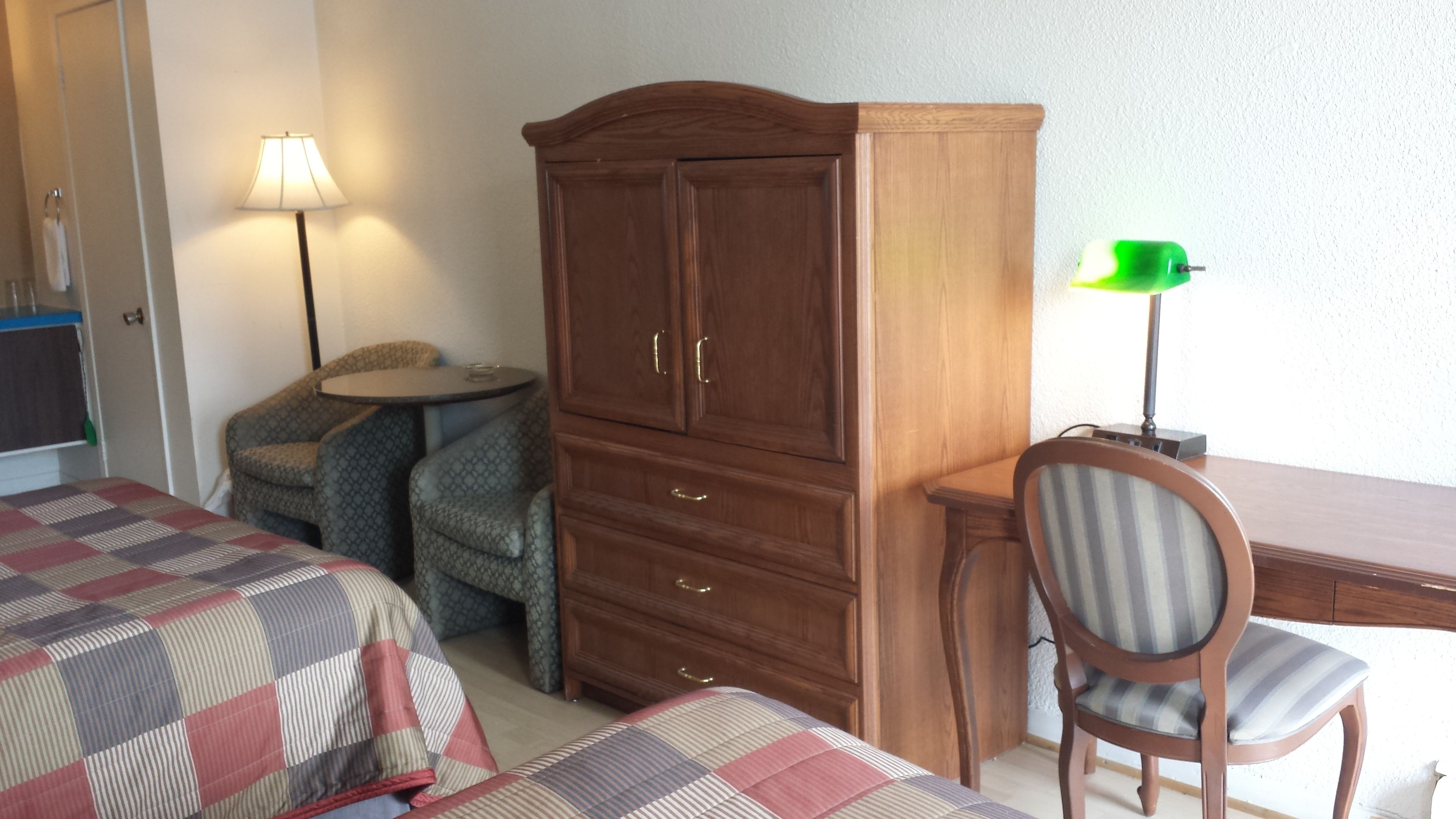 A hotel room with two beds, a wooden wardrobe, a small round table, a chair, and a desk with a lamp.