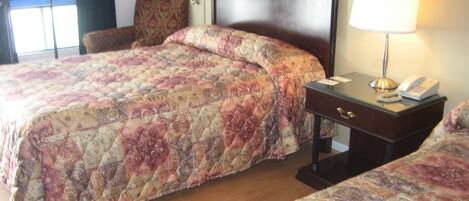 A hotel room with a large bed, a smaller bed, a nightstand with a lamp, and a chair.