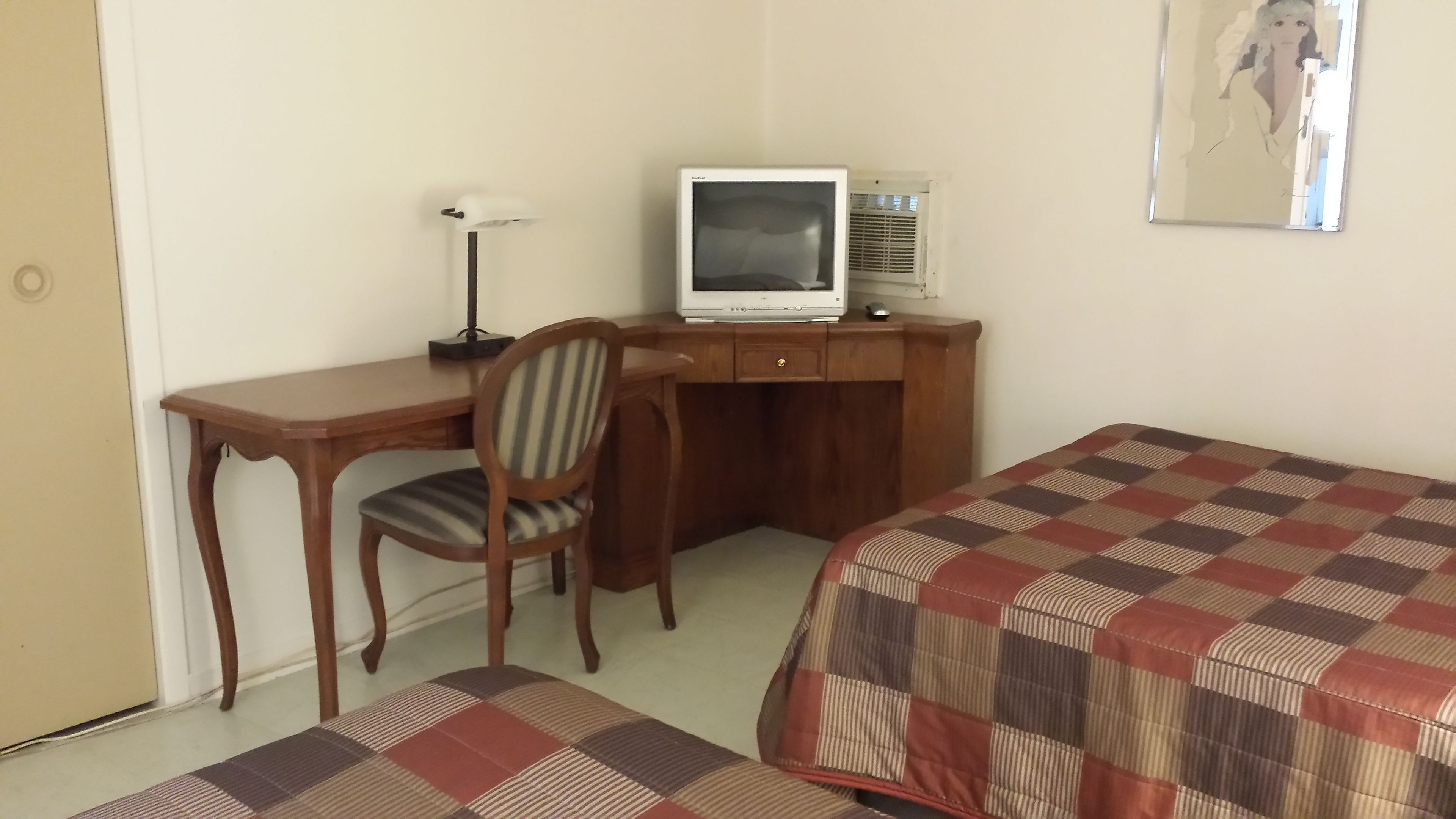 Una habitación con escritorio de madera, una silla, televisor, aire acondicionado y una cama con colcha a cuadros.
