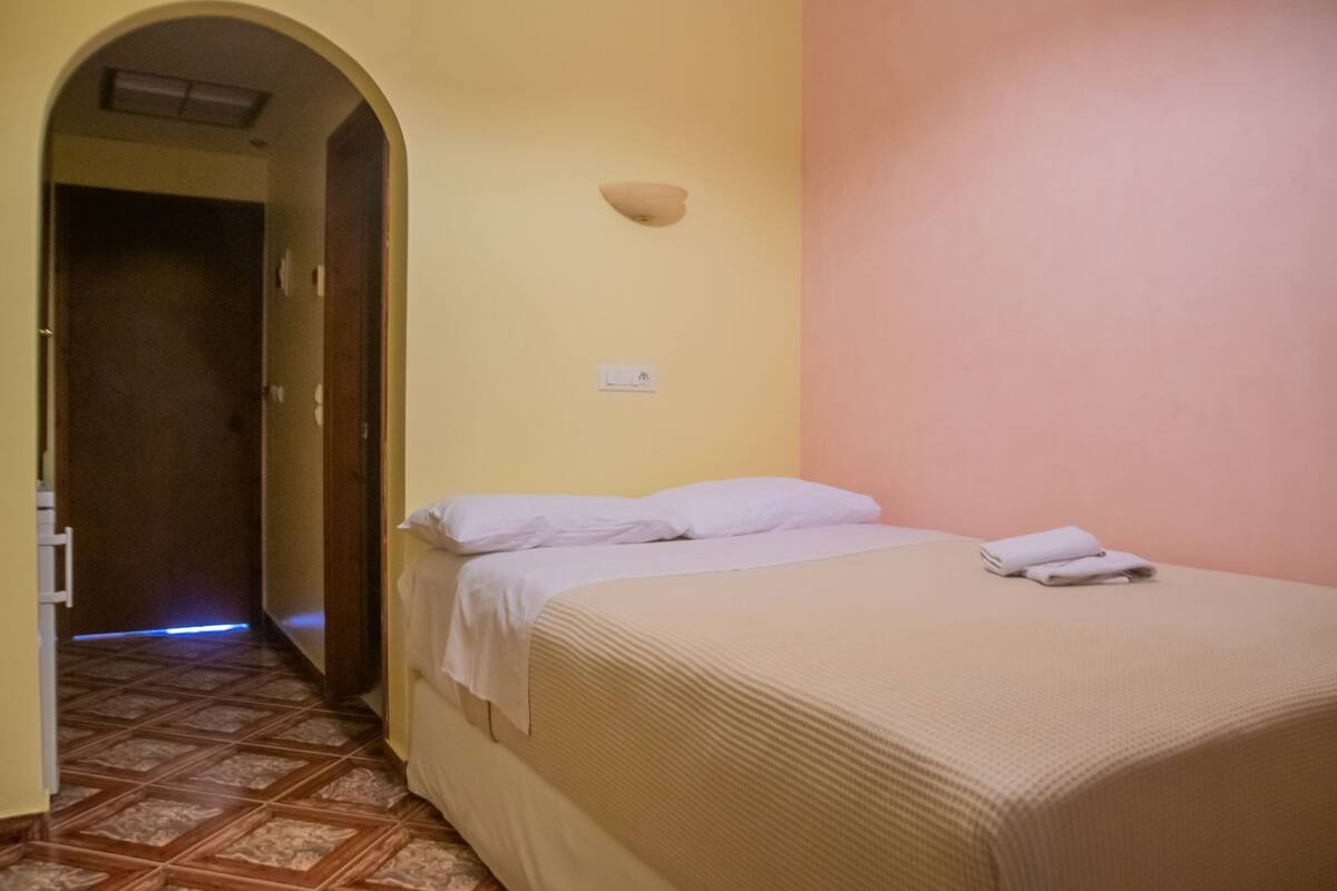 A room with a bed, a wall socket, and a door leading to another room.